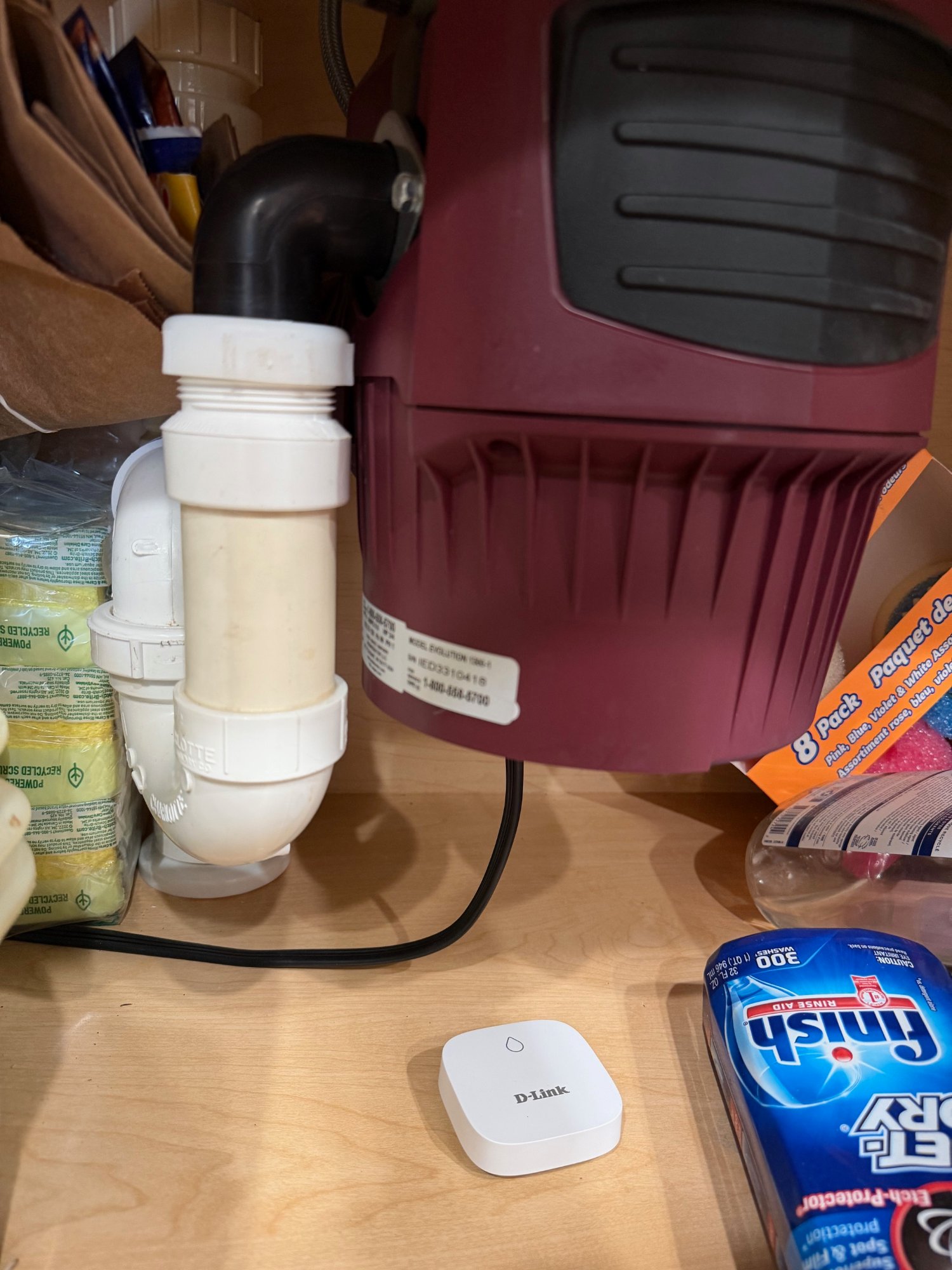 A white D-Link water leak sensor pod sitting on the shelf under a kitchen sink, next to drain pipes and a garbage disposal unit, monitoring for water leaks