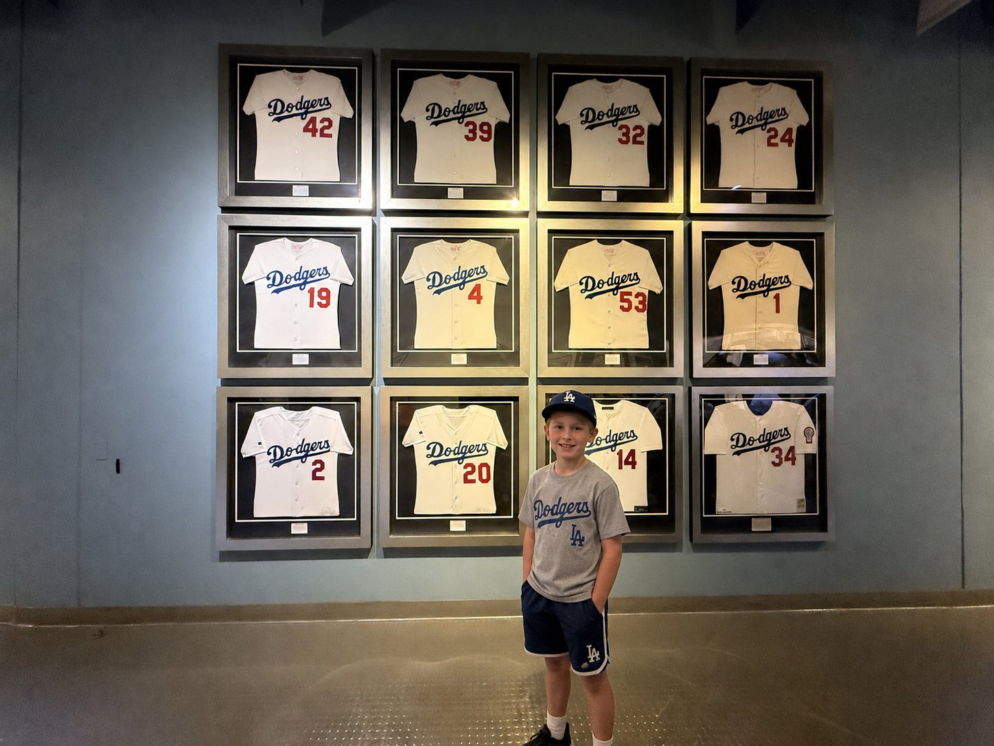 A smiling young boy wearing a Dodgers T-shirt and cap stands in front of a wall of framed Los Angeles Dodgers retired-number jerseys at Dodger Stadium.