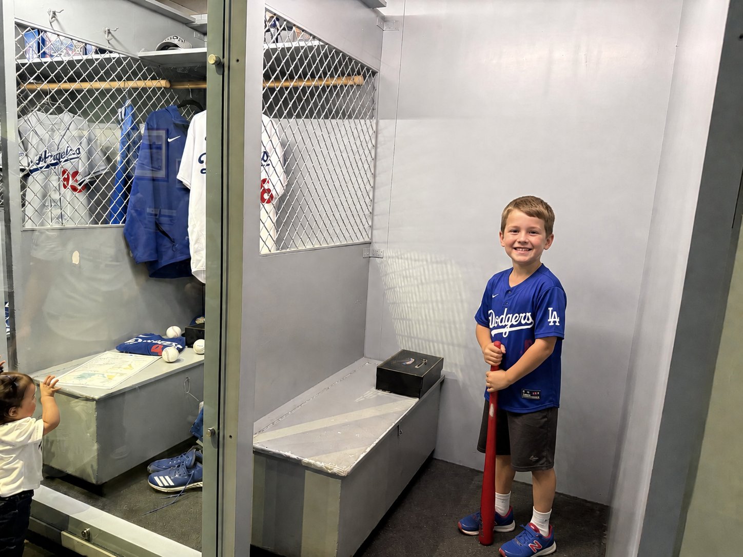 A smiling young boy in a blue Dodgers jersey holds a red bat inside a replica locker room exhibit at Dodger Stadium.