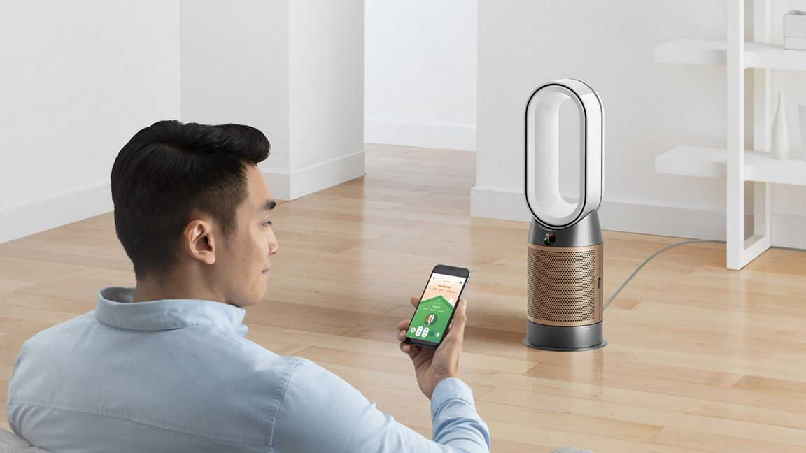 A man sits on a sofa checking his home’s air quality on the MyDyson phone app while a Dyson Purifier Hot+Cool stands across the room.